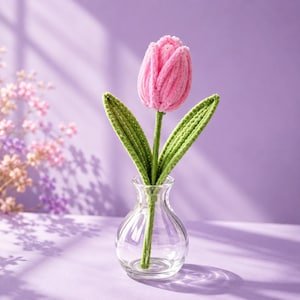 May include: A single pink crocheted tulip flower with two green crocheted leaves on a stem, placed inside a small, clear glass vase. The flower features a soft, textured yarn construction with visible stitching throughout the petals and leaves.