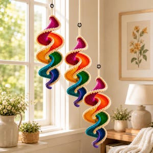 May include: Three hanging spiral ornaments featuring a vibrant rainbow gradient of magenta, orange, yellow, green, and blue yarn. Each piece is crocheted with a cream-colored border and suspended from a thin, light-colored rope with a black ring.