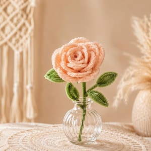 May include: A peach-colored crocheted rose with a textured, layered petal design and green crocheted leaves on a stem. The flower is displayed in a small, clear, ribbed glass vase with a narrow neck and a rounded base, standing upright.