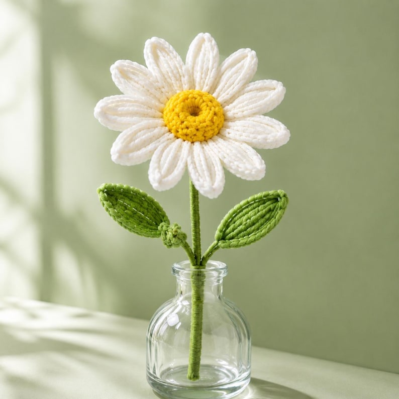 May include: A handcrafted crochet daisy flower with white petals and a bright yellow center, attached to a green stem with two green leaves. The flower is displayed in a clear, small glass bottle with a rounded base and a narrow neck.