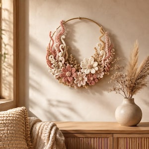 Può includere: Una decorazione da parete circolare con un cerchio in metallo dorato decorato con fiori all'uncinetto e viticci testurizzati a spirale in tonalità rosa polvere, crema e beige. La composizione floreale è disposta lungo la metà inferiore.