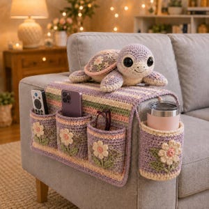 May include: A crochet sofa armrest organizer in pastel stripes with four pockets, each featuring a white flower applique. A crochet turtle plush with a floral shell sits on top. The pockets hold a remote, a smartphone, glasses, and a tumbler.
