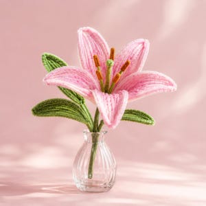 May include: A handcrafted crochet lily flower with six light pink petals, a central green pistil, and brown-tipped stamens. The flower features several textured green leaves and is displayed in a small, clear, ribbed glass vase.