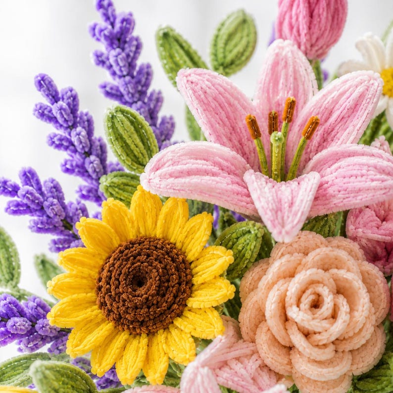 May include: A handcrafted crochet flower bouquet featuring a prominent pink lily with brown stamens, a bright yellow sunflower with a dark brown center, a peach-colored rose, and several sprigs of purple lavender, all accented by textured green leaves.