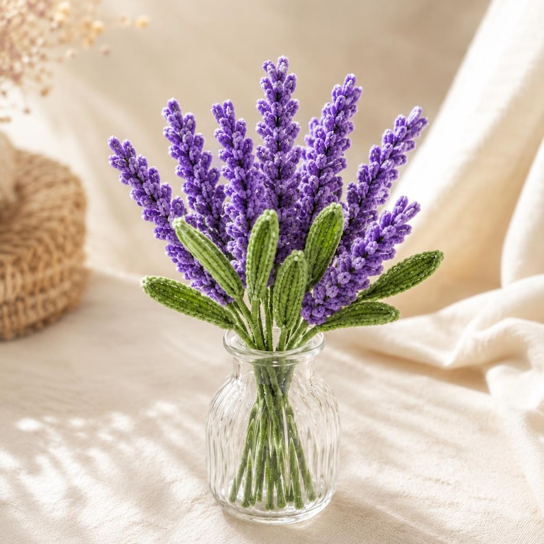 May include: A bouquet of crocheted lavender stems with vibrant purple flower heads and textured green leaves. The stems are arranged in a clear, ribbed glass vase, showcasing the intricate yarn work and detailed stitching of each individual flower.
