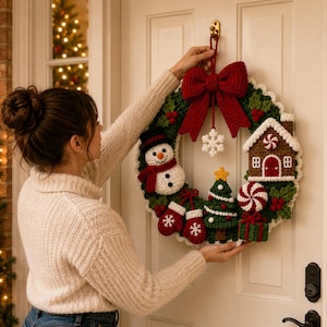 May include: A round Christmas wreath made of green crocheted yarn, featuring a large red bow at the top, a white snowman, a brown gingerbread house, a green Christmas tree, red mittens, a peppermint candy, and a green gift box.