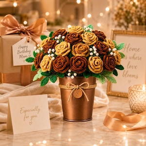 Pode incluir: Um bouquet de rosas com textura aveludada em tons de castanho e dourado, acentuado com pequenas flores brancas e folhas verdes, disposto num balde de bronze metálico. O balde apresenta um laço de corda e uma etiqueta circular com a letra E.