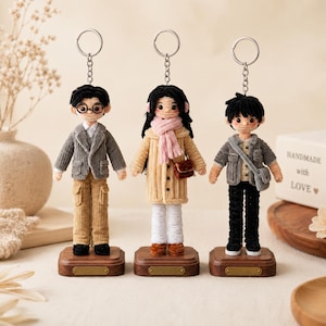 May include: Three handmade crochet dolls attached to silver keychains, each standing on a small rectangular wooden base. The dolls feature detailed yarn clothing, including jackets, scarves, and trousers, with distinct hairstyles and facial features.