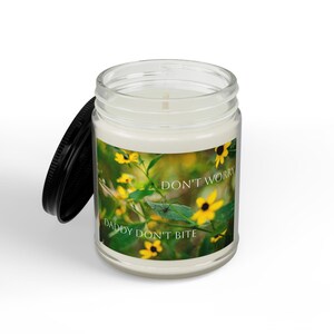 May include: A clear glass candle jar with a black lid, featuring a label with a photograph of yellow flowers and a small spider on a green leaf. The text on the label reads, "DON'T WORRY DADDY DON'T BITE" in white, capitalized, sans-serif lettering.