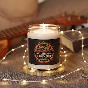 Op de afbeelding: Een witte geurkaars in een doorzichtige glazen pot met een zwart etiket voorzien van een vlammenafbeelding en de tekst: Love is a burning thing & it makes a fiery ring, bound by wild desire, I fell into a ring of fire. De kaars staat op een houten onderzetter.