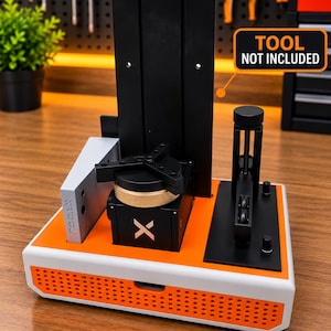 以下が含まれることがあります： 白とオレンジの卓上マシン。黒い垂直プレート、金色のダイヤルとXロゴが付いた中央の黒いブロック、TOOL.Xとラベル付けされた銀色の側面コンポーネント、黒い機械アームを備えています。オレンジ色の看板にはTOOL NOT INCLUDEDと書かれています。