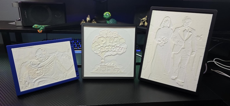 May include: Three framed 3D-printed white relief plaques. The left plaque is rectangular with a blue frame showing a skeleton. The centre square plaque has a black frame and the text The Pea Factory. The right plaque is rectangular with a black frame.