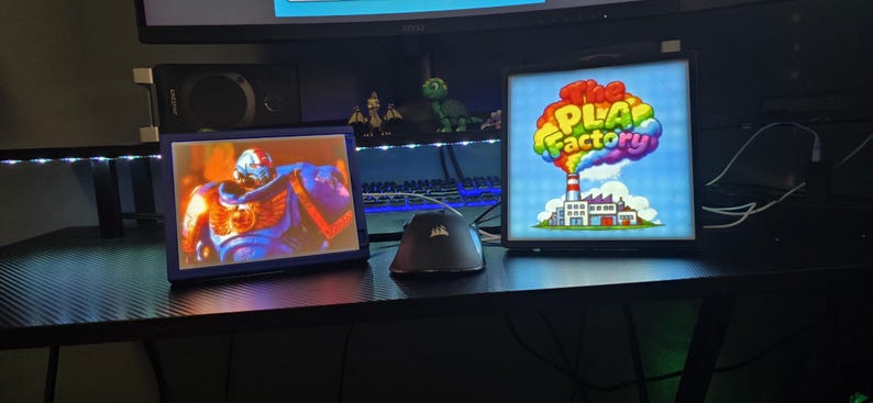May include: Two rectangular light boxes with black frames. The left box displays a blue armoured figure with gold accents. The right box features a factory illustration with a rainbow cloud and the text The PLA Factory in colourful letters.
