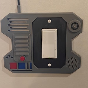 May include: A grey, rectangular wall-mounted light switch cover plate featuring a sci-fi inspired design with a vertical antenna, a circular speaker grille, and decorative red and blue buttons. A white Leviton rocker light switch is centred.