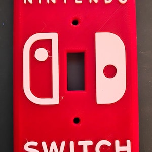 Pode incluir: Uma placa de parede retangular vermelha para interruptor de luz com o texto branco NINTENDO no topo e SWITCH na parte inferior. O centro exibe um gráfico branco de dois componentes de controle de jogo com um recorte retangular.