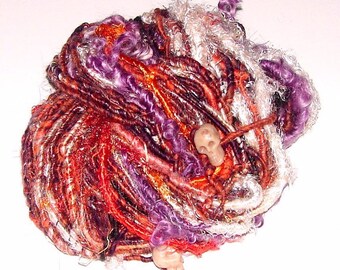 May include: A skein of hand-spun yarn in shades of purple, orange, red, and white. The yarn is arranged in a loose ball with two small skull-shaped beads.