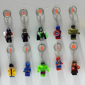 May include: A collection of fourteen metal keychains featuring various plastic superhero minifigures, each labeled with an orange letter from A to N. The figures include characters in red, blue, green, yellow, and brown outfits with accessories.