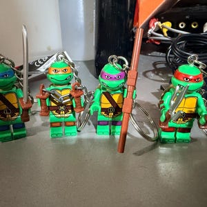 May include: Four Teenage Mutant Ninja Turtles character keychains, each featuring a green plastic figure with a yellow chest plate, a colored mask, and a unique weapon accessory. The figures include blue, orange, purple, and red masks with metal rings.