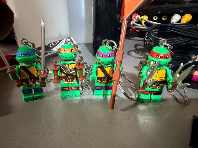 May include: Four Teenage Mutant Ninja Turtles character keychains, each featuring a green plastic figure with a yellow chest plate, a colored mask, and a unique weapon accessory. The figures include blue, orange, purple, and red masks with metal rings.