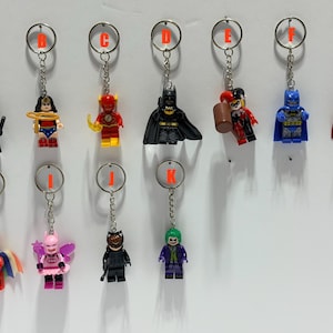 May include: A collection of eleven metal keychains featuring various plastic superhero and villain minifigures, each labeled with a red letter from A to K. The figures include characters in red, black, blue, and multicolored outfits with accessories.