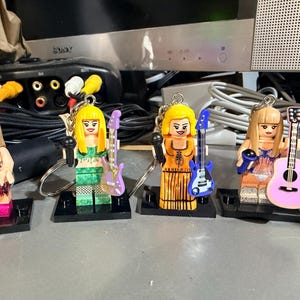 May include: Four plastic minifigure keychains featuring female characters with blonde hair, each holding a miniature microphone and a musical instrument. The figures wear distinct outfits including fringe, patterned, and solid dresses, attached to metal rings.