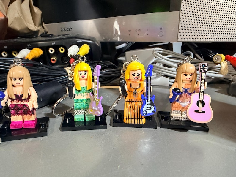 May include: Four plastic minifigure keychains featuring female characters with blonde hair, each holding a miniature microphone and a musical instrument. The figures wear distinct outfits including fringe, patterned, and solid dresses, attached to metal rings.