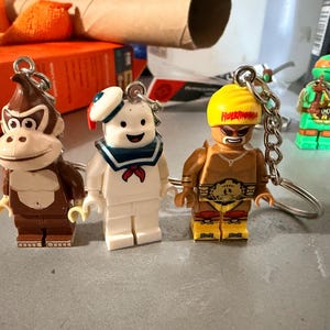 May include: Three character keychains featuring a brown gorilla, a white marshmallow-like figure with a blue sailor collar, and a tan wrestler with a yellow headband labeled Hulkamania, each attached to a silver metal ring and chain.