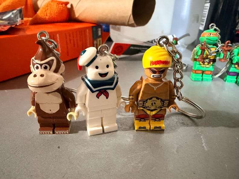 May include: Three character keychains featuring a brown gorilla, a white marshmallow-like figure with a blue sailor collar, and a tan wrestler with a yellow headband labeled Hulkamania, each attached to a silver metal ring and chain.