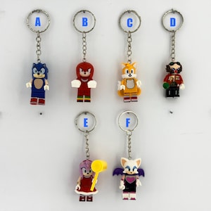 May include: Six metal keychains featuring plastic character figures labeled A through F. The figures include a blue hedgehog, a red echidna, a yellow fox, a mustachioed villain, a pink hedgehog with a yellow hammer, and a white bat with purple wings.