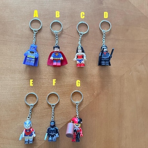 May include: Seven metal keychains featuring miniature plastic superhero and villain figures labeled A through G. The figures include Batman, Superman, Wonder Woman, Robin, a character with a silver helmet, a black armored figure, and Harley Quinn.