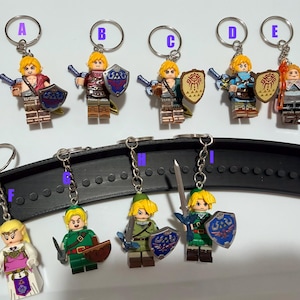 May include: A collection of nine plastic minifigure keychains labeled A through I, each featuring a character with blonde hair, pointed ears, and various accessories including swords and shields, attached to silver metal key rings and chains.