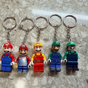 May include: Five character keychains featuring plastic figures of Mario and Luigi in various outfits. Each figure is attached to a silver metal chain and ring. The figures include two Marios in blue overalls, one Mario in yellow and red, and two Luigis.