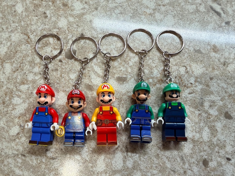May include: Five character keychains featuring plastic figures of Mario and Luigi in various outfits. Each figure is attached to a silver metal chain and ring. The figures include two Marios in blue overalls, one Mario in yellow and red, and two Luigis.