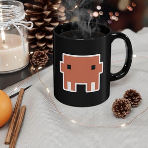 May include: A black ceramic mug featuring a stylised, pixelated orange creature graphic with two square black eyes and two white rectangular teeth. The mug has a smooth finish, a curved handle, and is filled with a steaming hot dark liquid.