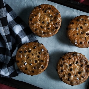 May include: Four round, golden-brown chocolate chip cookies arranged on a rectangular baking sheet lined with white parchment paper, accompanied by a folded black and white buffalo check cloth napkin resting on the side of the tray.