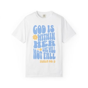 Op de afbeelding: Een wit t-shirt met korte mouwen met de blauwe tekst "GOD IS WITHIN HER SHE WILL NOT FALL" in een golvend lettertype. Twee kleine gele bloempictogrammen zijn naast de tekst geplaatst en de woorden "PSALM 46:5" staan in het geel onderaan.