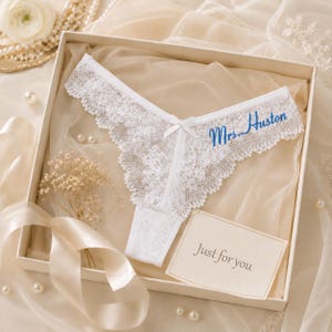 Puede incluir: Una tanga de encaje blanco con un pequeño lazo central y el texto bordado en azul Mrs. Huston. La prenda se presenta en una caja junto a una tarjeta con el texto impreso Just for you, rodeada de cintas decorativas y perlas.