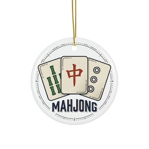 May include: A round white ceramic ornament featuring a graphic illustration of three mahjong tiles. The center tile displays a red Chinese character, flanked by green bamboo and circular patterns. The word MAHJONG is printed in dark blue below the tiles.