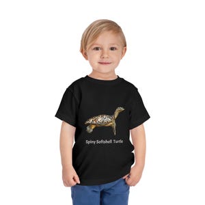 May include: A black short-sleeve t-shirt featuring a realistic graphic of a spiny softshell turtle in the center. Below the turtle illustration, the text Spiny Softshell Turtle is printed in a clean, white, sans-serif font on the front of the shirt.