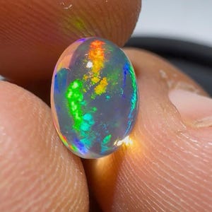 Puede incluir: Una piedra preciosa de ópalo negro de forma ovalada con un cuerpo oscuro translúcido y destellos vibrantes de verde neón, naranja brillante y azul eléctrico en toda la piedra, con una superficie de cabujón pulida, lisa y redondeada.