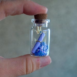May include: A small clear glass bottle with a cork stopper containing a rolled purple paper scroll, a gold-coloured letter M charm, and a collection of tiny blue and purple flower-shaped confetti pieces resting at the bottom of the glass container.