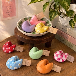May include: A collection of colourful, hand-painted fortune cookie-shaped ornaments made of clay. Each piece features unique patterns like daisies, hearts, or checkers and includes a small paper tag with handwritten messages like You Got This.