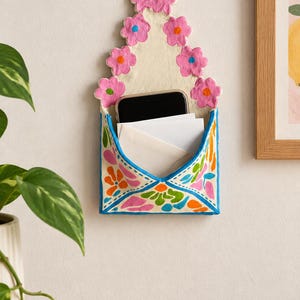 May include: A wall-mounted envelope-shaped organiser made of off-white material, featuring a top section decorated with pink flowers and a lower pocket painted with colourful floral patterns in blue, orange, and green, holding a phone and mail.
