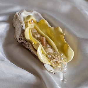 Könnte beinhalten: Eine gelbe Muschel aus Kunstharz, gefüllt mit Muscheln, Perlen und einem Seestern. Die Muschel liegt auf einer weißen Stoffoberfläche.