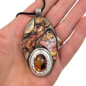 Puede incluir: Un colgante ovalado de plata con una piedra marrón y naranja engastada en el centro. El colgante está rodeado de un mosaico de piezas de cobre y oro.