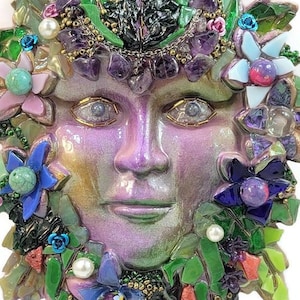 Puede incluir: Una placa de pared con un rostro esculpido con un acabado nacarado en morado y verde, rodeado por un mosaico de hojas de vidrio verde, piedras de amatista moradas, cuentas de perlas blancas y varios acentos florales de vidrio y metal.