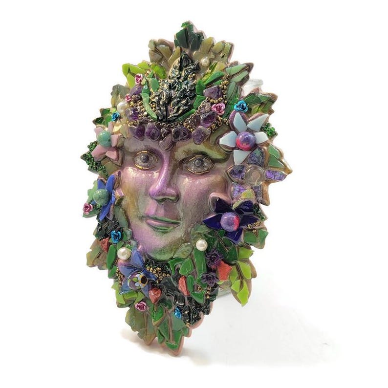 Puede incluir: Una m&aacute;scara decorativa de pared con un rostro humano central de acabado iridiscente en tonos morados y verdes, rodeado por un denso mosaico de hojas de vidrio verde, peque&ntilde;as flores coloridas, piedras de amatista y perlas blancas.