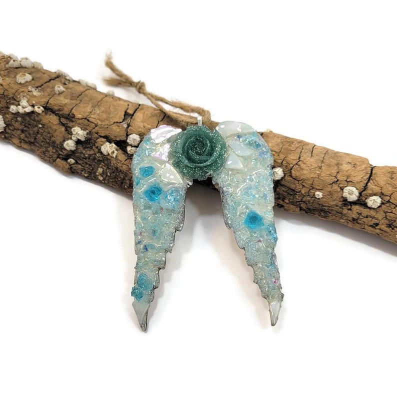 May include: A handcrafted angel wing ornament with a teal rose center. The wings are light blue with iridescent accents and turquoise details. The ornament is suspended by a natural twine cord and rests on a piece of driftwood.