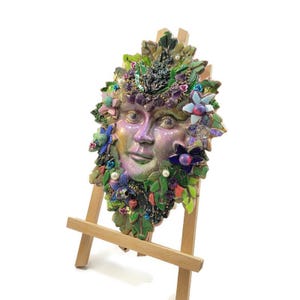 Puede incluir: Una placa decorativa de pared con un rostro central de color p&uacute;rpura iridiscente rodeado por un mosaico de hojas verdes, piedras de amatista moradas, peque&ntilde;as perlas blancas y varios acentos florales coloridos, exhibida sobre un caballete de madera clara.