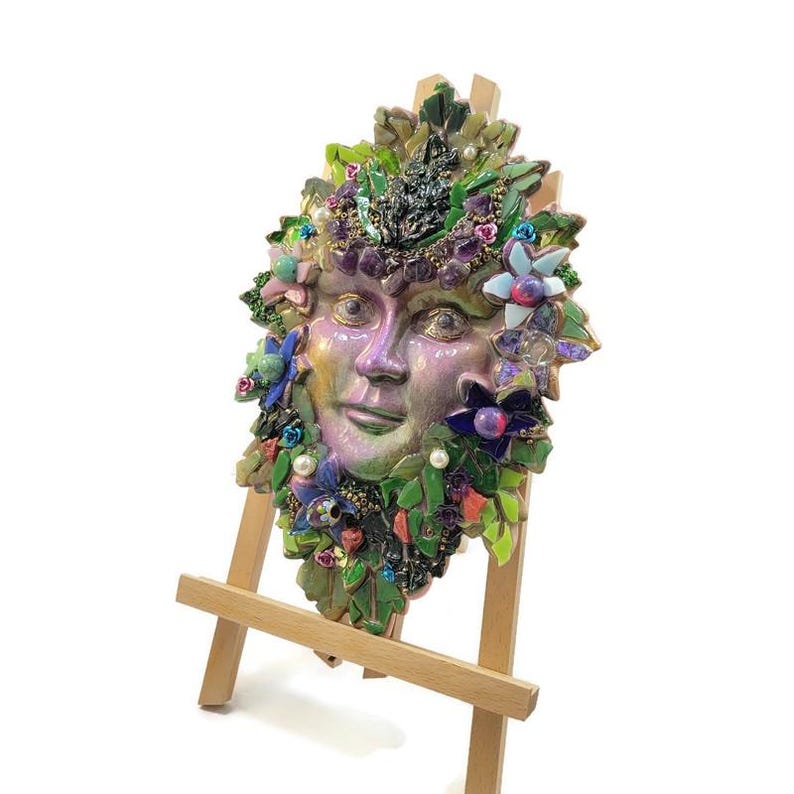 Puede incluir: Una placa decorativa de pared con un rostro central de color p&uacute;rpura iridiscente rodeado por un mosaico de hojas verdes, piedras de amatista moradas, peque&ntilde;as perlas blancas y varios acentos florales coloridos, exhibida sobre un caballete de madera clara.